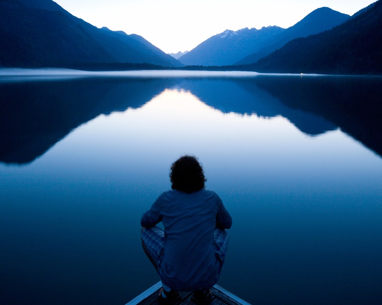 Man sitting by lake admiring serene view of mountains reflection in the water
